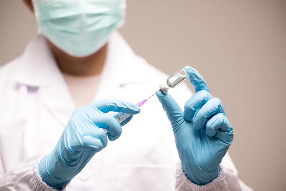 Médico com luva azul segurando agulha e frasco de vacina.
