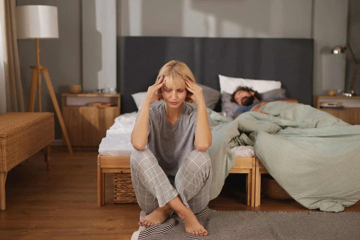 Mulher sentada aos pés da cama, preocupada enquanto o marido dorme ao fundo, podendo demonstrar como o desconforto e a dor causados pelo líquen escleroso podem afetar a intimidade, o desejo e a vida sexual do casal.